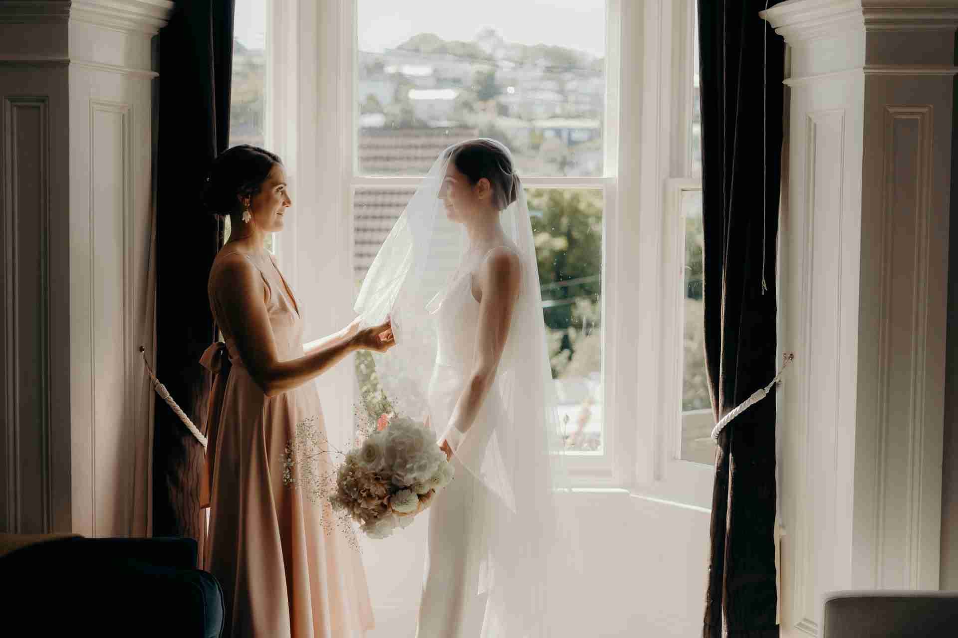 A bridesmaid fixes the bride's veil by a window