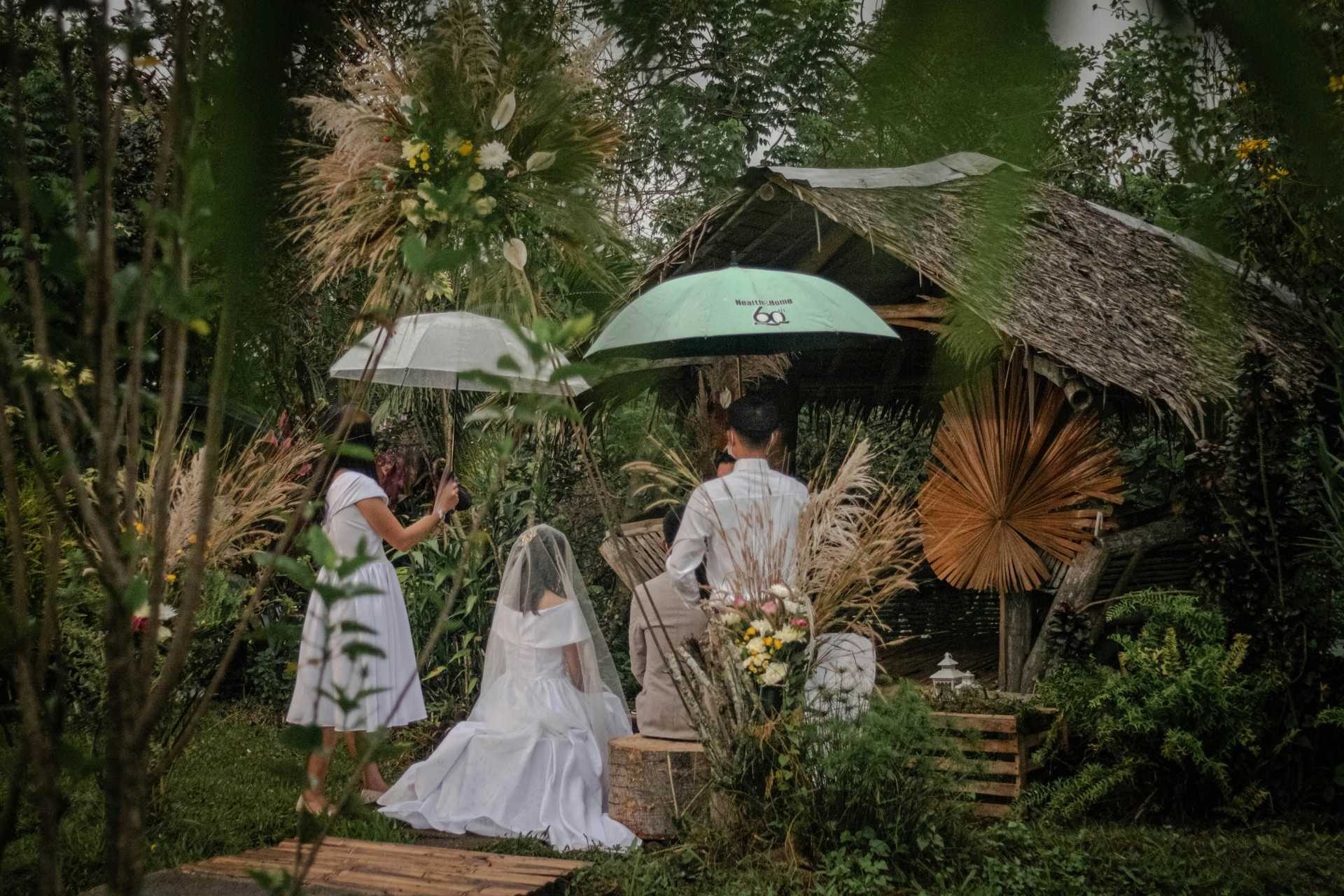 A small outdoor wedding where a couple sits under umbrellas in a garden setting.