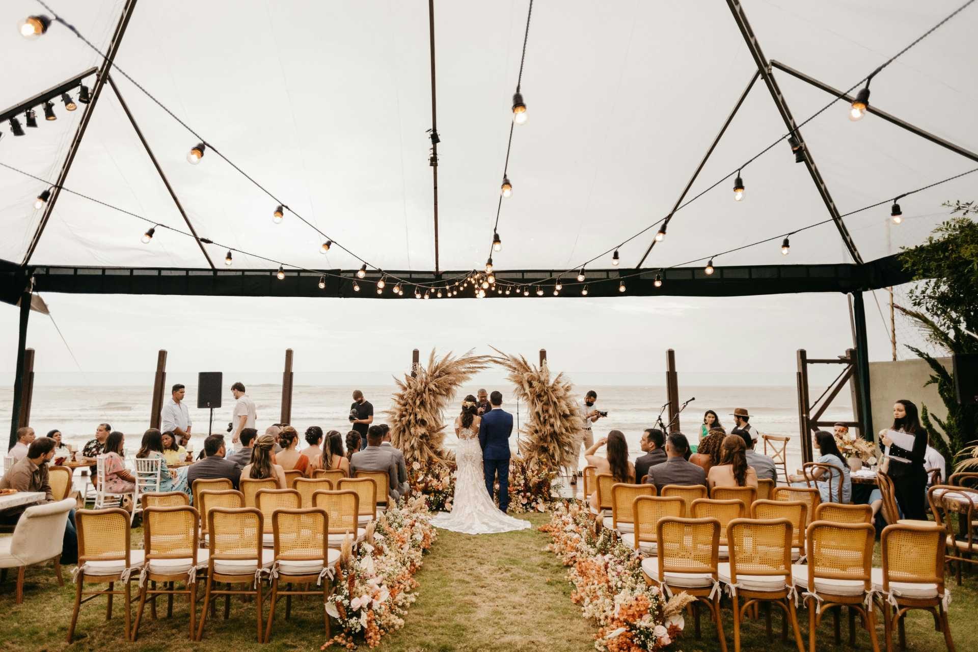 A small outdoor wedding ceremony by the beach, with a couple standing at the altar framed by pampas grass arrangements, surrounded by a few guests